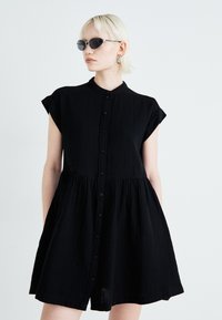 Rochie neagră cu mâneci scurte, cu închidere prin nasturi, talie strânsă și croială lejeră. Fabricată dintr-un material ușor, cu o textură subtilă.