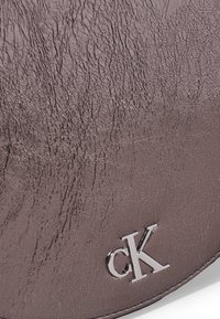 Portafoglio in pelle marrone con texture, dotato di logo cK argentato, caratterizzato da una finitura liscia e pieghe sottili per un aspetto tattile.