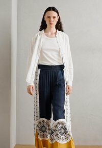 White lightweight jacket over a fitted white top, paired with navy blue loose trousers featuring a circular patterned hem in white and gold.