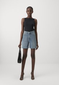 Zwarte mouwloze top, hoog aangesneden lichtdenim shorts met uitgerafelde zoom, zilveren haksandalen, met een zwarte schoudertas.