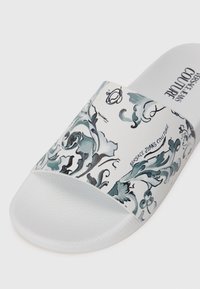 White slide sandal with a decorative gray floral pattern and "Versace Jeans Couture" text. Textured white outsole for grip.