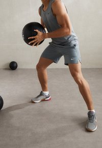 Grauer sportlicher Tanktop und Shorts, hält einen schwarzen Medizinball. Sneakers mit grauen, roten und tanfarbigen Akzenten auf einem glatten, neutralen Boden.