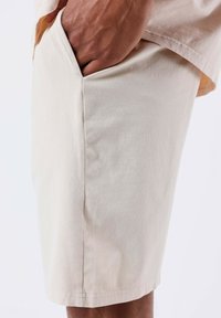 Pantaloni corti beige chiaro realizzati in tessuto di cotone con una texture liscia. Presentano una tasca laterale e cuciture pulite lungo l'orlo.