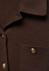 Cardigan en maille côtelée marron foncé avec col replié, poche poitrine boutonnée et trois boutons ronds marron visibles.