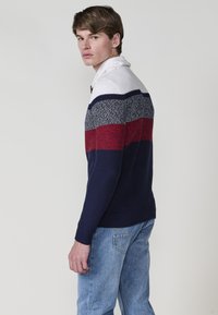 Modelo masculino con un suéter de bloques de color en azul marino, rojo y blanco, de pie contra un fondo simple, mirando por encima del hombro.
