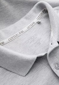 Cuello de polo de algodón gris con una tela texturizada, que presenta una etiqueta grabada con "línea de algodón" y "ombre" dentro del escote.