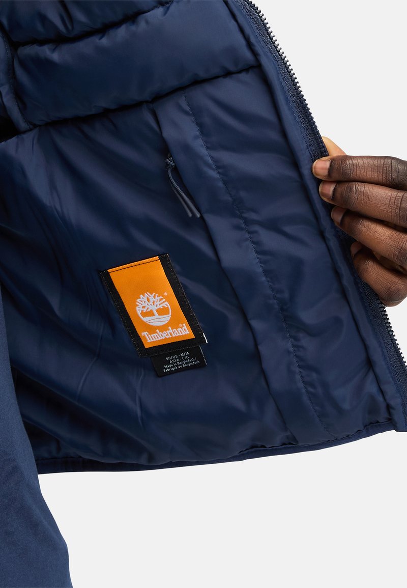 Interno di un giubbotto imbottito blu navy con una texture trapuntata, caratterizzato da un'etichetta con il logo Timberland arancione e nero, con un dettaglio della tasca con zip.