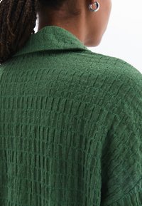 Camicia di un verde scuro con una texture a griglia e un colletto ampio, realizzata in un tessuto morbido e leggero e con una vestibilità leggermente ampia.