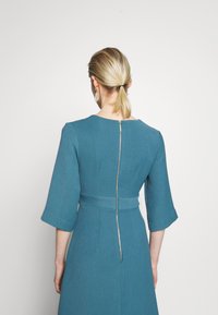 Une robe bleue avec un tissu texturé, présentant un corsage ajusté, des manches larges et une fermeture éclair dorée visible au dos.