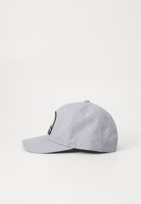Căciulă de baseball gri cu o rimă curbată, având un patch brodat pe partea stângă și orificii de ventilație în formă de ochi. Textură netedă.