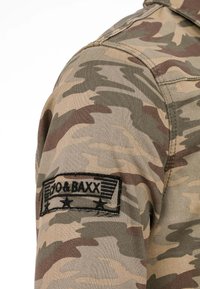 Kurtka camouflage z mieszanką zielonych, brązowych i beżowych wzorów. Zawiera czarną haftowaną naszywkę z napisem "DO & BAXX" na ramieniu.
