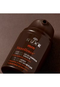 Fluide anti-âge NUXE Nuxellence pour hommes dans un flacon marron foncé avec un design incurvé, arborant un texte blanc et orange, texture lisse.
