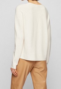 Pull en tricot couleur crème, coupe décontractée, épaules tombantes et texture côtelée. Associé à un pantalon marron clair, mettant en valeur une tenue décontractée.