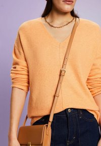 Jasnopomarańczowy sweter robiony na drutach z dekoltem w serek i podwiniętymi rękawami, połączony z ciemnoniebieskimi dżinsami i beżową torbą na ramię. Akcent w postaci łańcuszka.