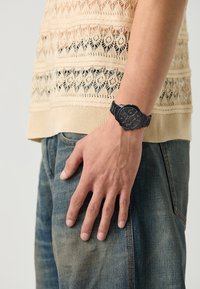 Svart klokke med rund urskive, teksturert skive og merkede subskiver, båret på et håndledd med en lysstrikket beige genser og blå jeans.