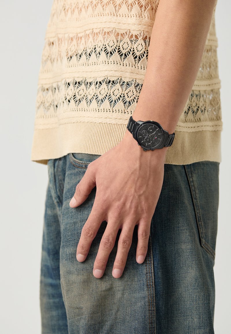 Svart klokke med rund urskive, teksturert skive og merkede subskiver, båret på et håndledd med en lysstrikket beige genser og blå jeans.