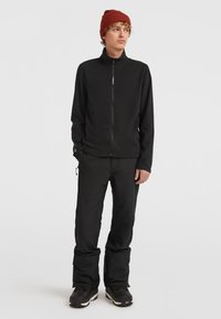 Sort zip-up jakke med høj krave, parret med sorte bukser og sorte snestøvler. En rød ribbet hue fuldender outfittet.