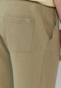 Detailansicht einer beige Hose mit sichtbaren Nähten und kleinem Logotag an der Gesäßtasche, die einen seitlichen Schnitt und einen Teil der Hand zeigt.