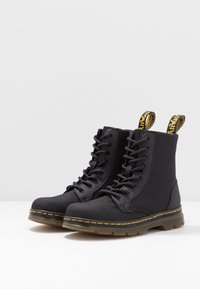 Black ankle boots made of textured fabric, featuring a lace-up design, yellow stitching, and pull tabs on the shaft for easy wear.