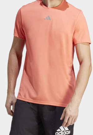 Muž v korálovém sportovním tričku s krátkým rukávem a černých šortkách Adidas, stojící s rukama volně podél těla před neutrálním pozadím.