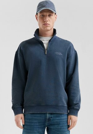 Sweatshirt bleu marine avec un col montant et une fermeture éclair, arborant un texte blanc sur la poitrine. Porté avec une casquette en denim assortie et un jean clair.