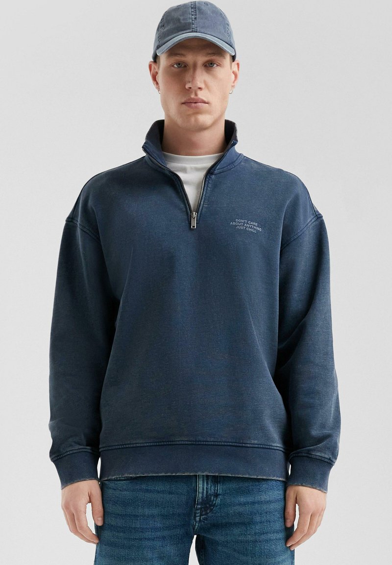 Marineblauer Sweatshirt mit hohem Kragen und Reißverschluss, versehen mit weißem Text auf der Brust. Getragen mit einer passenden Denim-Kappe und hellen Jeans.