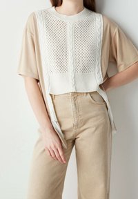 Top beige a maniche corte con un pannello lavorato a maglia bianco con dettagli a treccia e frange. Abbinato a pantaloni beige chiaro a vita alta.