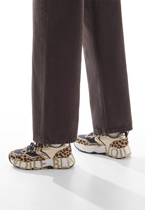 Sneaker con stampa leopardata e accenti neri e marroni, suola chunky e testo "VOILE" in evidenza sul lato, abbinata a pantaloni larghi marrone scuro.