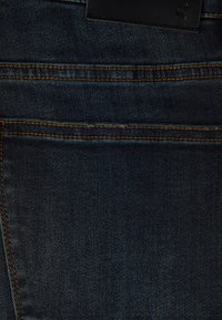 Mörkblå denimtyg med synlig vävstruktur, orange sömmar och en svart läderetikett högst upp. Klassisk jeansdesign.