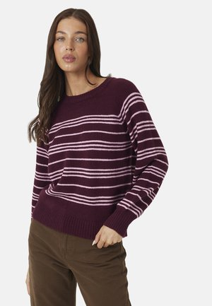 Frau mit langen braunen Haaren, die einen lila und pink gestreiften Pullover und braune Hose trägt, steht vor einem einfarbigen Hintergrund.