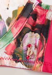 Nahaufnahme von floraler Stoff mit rosa, grünen und roten Tönen, detaillierter Naht und einem kleinen goldenen "b"-Anhänger, der mit rotem Faden befestigt ist.