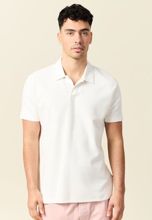 Junger Mann mit kurzen, lockigen schwarzen Haaren, trägt ein schlichtes weißes Polohemd mit kurzen Ärmeln und hellrosa Hose vor einem beigen Hintergrund.