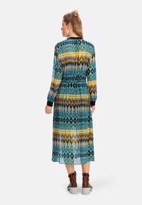 Maxikleid mit einem bunten Zickzackmuster in Blau, Gelb und Schwarz. Elastischer Taillenbund und lange Ärmel, kombiniert mit braunen Ankle Boots.