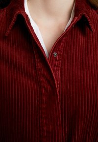 Bordeaux corduroy shirt met verticale ribstructuur, voorzien van een puntkraag, drukknoopsluiting en een gelaagd wit ondershirt dat eronder zichtbaar is.