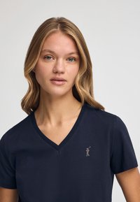 Marinblå kortärmad t-shirt med V-ringning i bomull, med en liten metallisk logodetalj på vänster bröst. Slät textur.