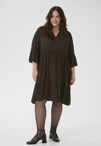 Rochie dintr-un brun închis, cu straturi, cu mâneci scurte, decolteu în V și model cu dungi metalice. Modelul poartă pantofi cu toc din piele lăcuită negri și colanți.