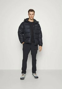 Černá péřová bunda s kapucí a zipovým uzávěrem, v kombinaci s černým tričkem a tmavými kalhotami. Doplněna šedými a oranžovými teniskami.