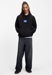 Felpa nera con cappuccio, patch logo blu, ampia tasca frontale e vestibilità leggermente ampia, abbinata a jeans denim scuri e scarpe nere.