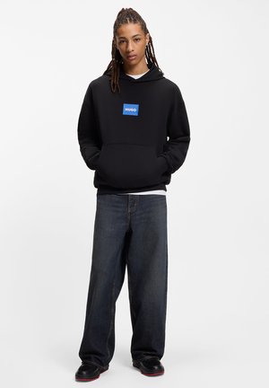 Black hoodie with a blue logo patch, large front pocket, and a slightly loose fit, paired with dark denim jeans and black shoes.