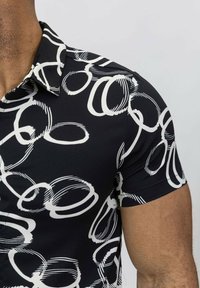 Chemise noire à manches courtes avec un motif circulaire abstrait blanc. Dotée d'un col et d'une texture de tissu lisse et extensible.