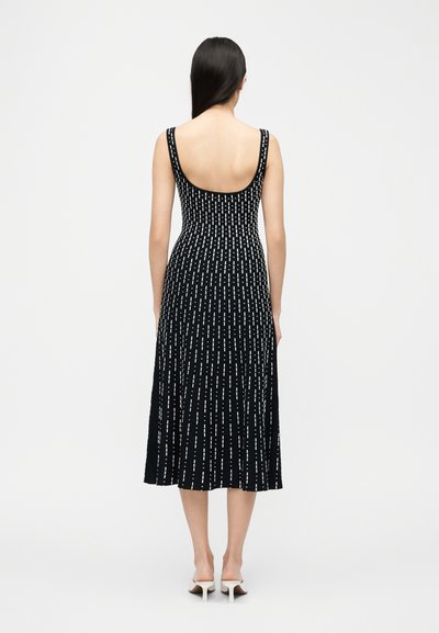 Γυναίκα με μακριά μαύρα μαλλιά, γυρισμένη με την πλάτη, φοράει μαύρο μεσαίο φόρεμα με λευκές κάθετες γραμμές και λευκές σανδάλια με τακούνι.