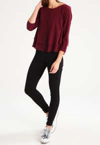 Haut superposé bordeaux à manches trois-quarts, associé à un jean skinny noir et des chaussures à plateforme argentées. Tissu lisse, coupe décontractée.