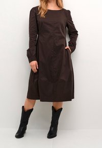 Ruskea, puolipitkä mekko, jossa on pitkät hihat, pyöreä pääntie ja sivutaskut. Yhdistetty mustiin cowboy-kengiin, joissa on koristeellinen ommel.