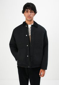 Young man wearing a black buttoned jacket with pockets over a plaid zip-up sweater and black pants, standing against a white background.