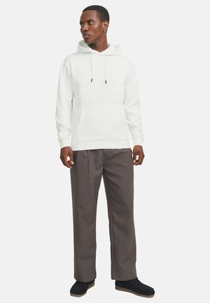 Homme debout, portant un sweat à capuche blanc, un pantalon gris et des chaussures noires, regardant vers sa gauche sur un fond uni.