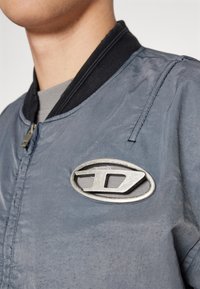 Veste bomber grise avec un col noir côtelé, présentant un logo ovale métallique sur la poitrine gauche et une texture légèrement froissée.