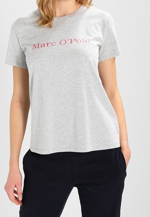 Kvinde iført en lysegrå T-shirt med korte ærmer med teksten "Marc O'Polo" i rødt, kombineret med mørke bukser, stående med den ene hånd i lommen.