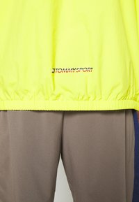 Lys gul vindjakke med elastisk kant, der har "TOMMYSPORT" logo i sort og rødt. Blødt, let materiale. Sammen med brune bukser.