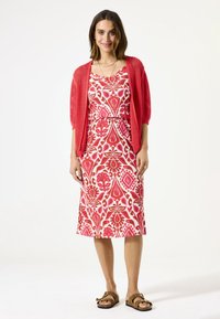 Robe rouge à motifs avec un cardigan en tricot rouge assorti, à manches courtes et coupe mi-longue. Sandales marron avec des accents en métal.