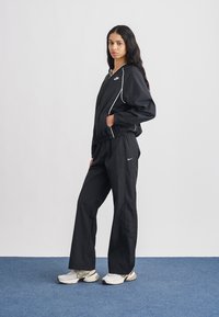 Fekete sportos melegítő könnyű anyagból, kontrasztos fehér szegélyekkel, laza szabással és mellrészen elhelyezett logóval díszítve.